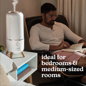 Man sitting in chair reading a book in his bedroom with the Homedics® Ultrasonic Humidifier on the table next to him. Ideal for bedrooms & medium-sized rooms