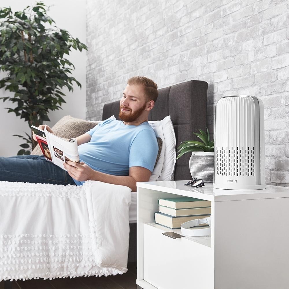 White | Man sits in bed next to the white Homedics TotalClean 4-in-1 Small Room Air Purifier