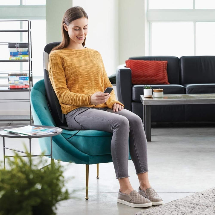 Woman looks at controller and relaxes in chair while enjoying a massage from the Homedics 8-Node Shiatsu Plus Massage Cushion with Soothing Heat