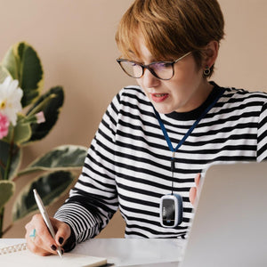 Woman writes at computer desk while wearing Homedics Premium Pulse Oximeter around neck