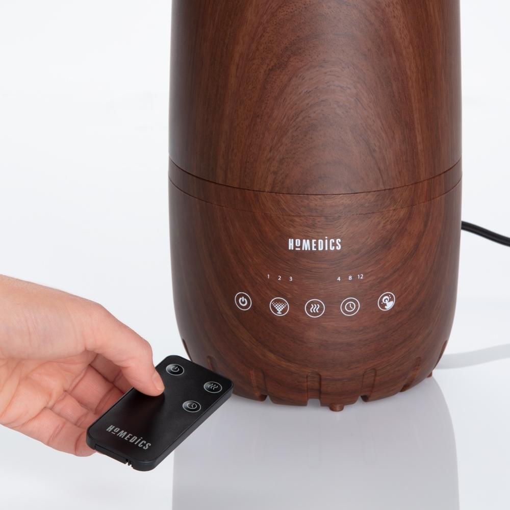 Hand holding remote control next to the Homedics TotalComfort 2-in-1 Warm and Cool Humidifier