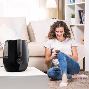 Woman sits next to couch looking at mobile phone screen with the Homedics TotalComfort Plus Ultrasonic Humidifier in the foreground